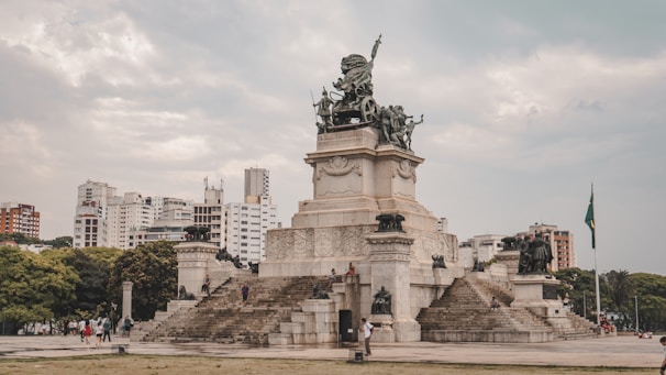 A large monument with detailed sculptures stands prominently in an urban park. The monument has a base with stairs leading up to a statue of a figure holding a weapon, surrounded by other figures and ornate carvings. Behind the monument, modern high-rise buildings stretch across the horizon under a slightly overcast sky. Trees and greenery surround the area, providing a contrast to the urban backdrop.