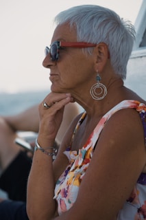 An older woman with short white hair is wearing sunglasses, large hoop earrings, and a sleeveless floral top. She is resting her chin on her hand, appearing to be deep in thought. There are blurred background elements that indicate she is outdoors, possibly on a boat near the water.