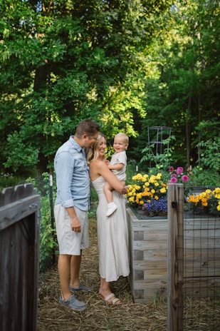 A family of three stands in a garden next to a raised wooden planter filled with vibrant flowers, including yellow and purple blossoms. The adults are casually dressed, and the child is being held by one of the adults. The background is filled with lush green foliage, creating a serene natural setting.