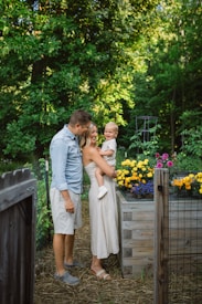 A family of three stands in a garden next to a raised wooden planter filled with vibrant flowers, including yellow and purple blossoms. The adults are casually dressed, and the child is being held by one of the adults. The background is filled with lush green foliage, creating a serene natural setting.