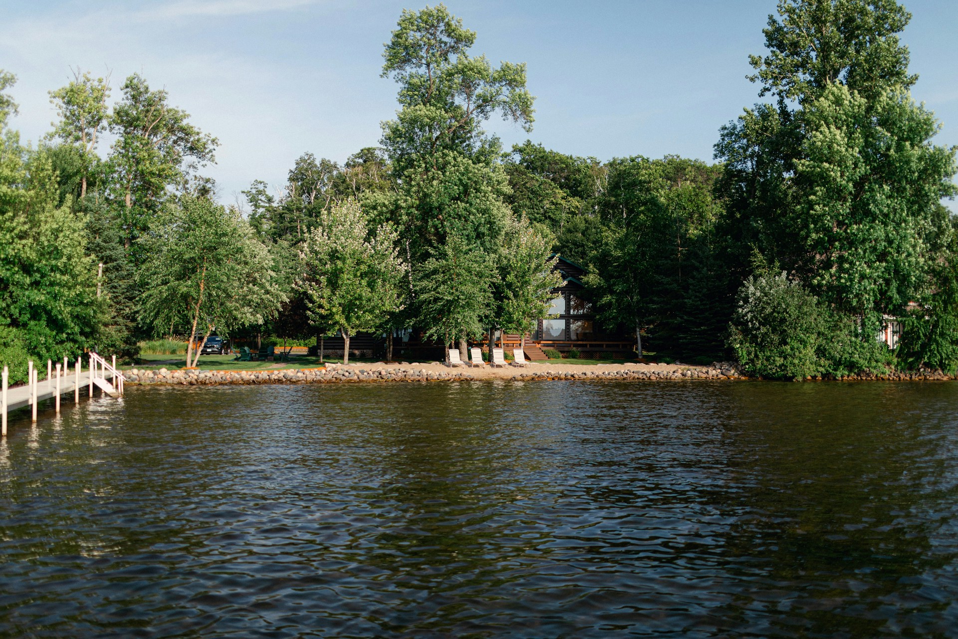 A peaceful lakeside scene with a dense border of tall, green trees surrounding a house. The calm water reflects the colors of the surroundings. A small dock extends into the lake, and there are chairs placed on the shore, suggesting a place for relaxation.