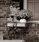 A rustic setup featuring a metal cart with decorative pumpkins and chrysanthemums placed in front of a brick wall. A sign saying 'Welcome Fall' hangs above the cart, decorated with illustrations of pumpkins and flowers.
