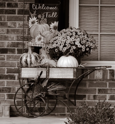 A rustic setup featuring a metal cart with decorative pumpkins and chrysanthemums placed in front of a brick wall. A sign saying 'Welcome Fall' hangs above the cart, decorated with illustrations of pumpkins and flowers.