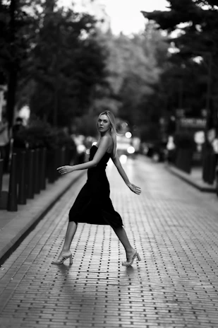 Elegant model wearing a flowing black dress with gold accents, walking through a softly lit urban evening scene.