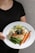 A person holding a plate with a gourmet meal that includes a grilled chicken breast topped with herbs, steamed green beans, roasted carrots, and a lemon wedge, all served over a creamy sauce.