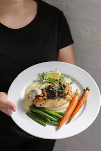 A person holding a plate with a gourmet meal that includes a grilled chicken breast topped with herbs, steamed green beans, roasted carrots, and a lemon wedge, all served over a creamy sauce.