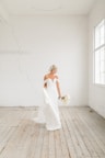 A bride in a strapless white wedding dress holding a bouquet stands in a bright, minimalist room with white brick walls and wooden flooring. The room is flooded with natural light from large windows.