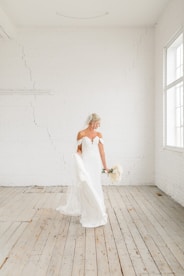 A bride in a strapless white wedding dress holding a bouquet stands in a bright, minimalist room with white brick walls and wooden flooring. The room is flooded with natural light from large windows.