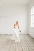 A bride in a strapless white wedding dress holding a bouquet stands in a bright, minimalist room with white brick walls and wooden flooring. The room is flooded with natural light from large windows.