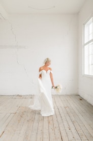 A bride in a strapless white wedding dress holding a bouquet stands in a bright, minimalist room with white brick walls and wooden flooring. The room is flooded with natural light from large windows.