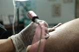 A close-up view of a person wearing gloves, holding a tattoo machine and working on a tattoo on someone's skin. The focus is on the hand and the tattoo machine, with a blurred background.
