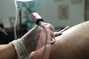 A close-up view of a person wearing gloves, holding a tattoo machine and working on a tattoo on someone's skin. The focus is on the hand and the tattoo machine, with a blurred background.