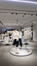 A modern clothing store interior featuring several mannequins dressed in casual summer apparel. The mannequins are positioned on a circular platform, each wearing different combinations of light-colored tops, shorts, and sandals. Surrounding the mannequins are clothing racks filled with various garments. The store is well-lit with overhead lights reflected in the smooth, tiled floor, giving the space a clean and organized look.