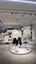A modern clothing store interior featuring several mannequins dressed in casual summer apparel. The mannequins are positioned on a circular platform, each wearing different combinations of light-colored tops, shorts, and sandals. Surrounding the mannequins are clothing racks filled with various garments. The store is well-lit with overhead lights reflected in the smooth, tiled floor, giving the space a clean and organized look.