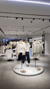 A modern clothing store interior featuring several mannequins dressed in casual summer apparel. The mannequins are positioned on a circular platform, each wearing different combinations of light-colored tops, shorts, and sandals. Surrounding the mannequins are clothing racks filled with various garments. The store is well-lit with overhead lights reflected in the smooth, tiled floor, giving the space a clean and organized look.