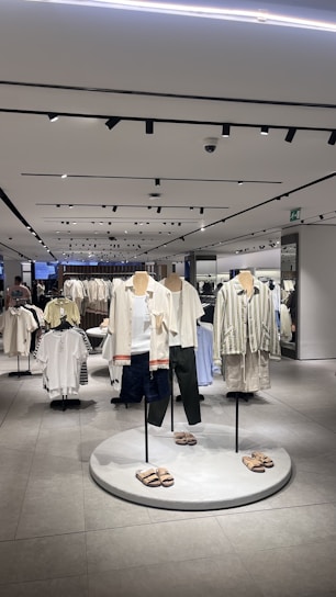 A modern clothing store interior featuring several mannequins dressed in casual summer apparel. The mannequins are positioned on a circular platform, each wearing different combinations of light-colored tops, shorts, and sandals. Surrounding the mannequins are clothing racks filled with various garments. The store is well-lit with overhead lights reflected in the smooth, tiled floor, giving the space a clean and organized look.
