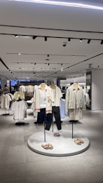 A modern clothing store interior featuring several mannequins dressed in casual summer apparel. The mannequins are positioned on a circular platform, each wearing different combinations of light-colored tops, shorts, and sandals. Surrounding the mannequins are clothing racks filled with various garments. The store is well-lit with overhead lights reflected in the smooth, tiled floor, giving the space a clean and organized look.
