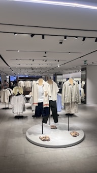 A modern clothing store interior featuring several mannequins dressed in casual summer apparel. The mannequins are positioned on a circular platform, each wearing different combinations of light-colored tops, shorts, and sandals. Surrounding the mannequins are clothing racks filled with various garments. The store is well-lit with overhead lights reflected in the smooth, tiled floor, giving the space a clean and organized look.