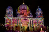 An architectural structure is illuminated with vibrant, multicolored lights projected onto its facade. The building features domes, columns, and ornate details that enhance the light display. Patterns include swirling designs and spots, creating a dynamic visual effect against the night sky.
