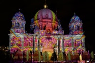 An architectural structure is illuminated with vibrant, multicolored lights projected onto its facade. The building features domes, columns, and ornate details that enhance the light display. Patterns include swirling designs and spots, creating a dynamic visual effect against the night sky.