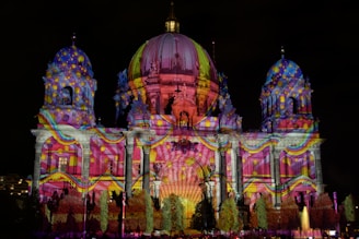 An architectural structure is illuminated with vibrant, multicolored lights projected onto its facade. The building features domes, columns, and ornate details that enhance the light display. Patterns include swirling designs and spots, creating a dynamic visual effect against the night sky.