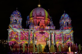 An architectural structure is illuminated with vibrant, multicolored lights projected onto its facade. The building features domes, columns, and ornate details that enhance the light display. Patterns include swirling designs and spots, creating a dynamic visual effect against the night sky.