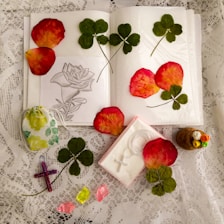 An arrangement on lace fabric featuring pressed clover leaves, rose petals, a floral sketch on paper, a small decorative pouch, a cross pendant box, a colorful beaded cross, crystals, and a small cork bottle with decorative faux flowers.