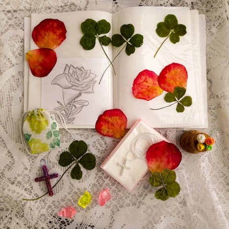 An arrangement on lace fabric featuring pressed clover leaves, rose petals, a floral sketch on paper, a small decorative pouch, a cross pendant box, a colorful beaded cross, crystals, and a small cork bottle with decorative faux flowers.