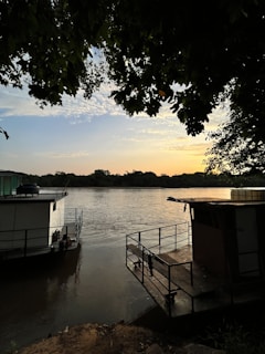 A serene riverside scene is captured with lush green tree branches overshadowing the view. Two houseboats are moored by the bank, gently floating on the calm water. The sky is beautifully painted with hues of orange and blue as the sun sets on the horizon.