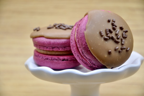 Two pink macarons with a light brown creamy topping and chocolate sprinkles are placed on a white ceramic pedestal. The background has a soft, blurred beige tone, creating a warm and inviting atmosphere.