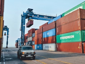 Stacks of shipping containers in various colors are arranged at a port. A large crane is lifting one of the containers. A vehicle is parked on the paved ground next to the containers.