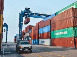 Stacks of shipping containers in various colors are arranged at a port. A large crane is lifting one of the containers. A vehicle is parked on the paved ground next to the containers.