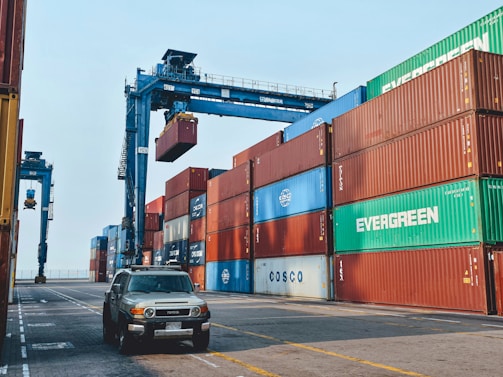 Stacks of shipping containers in various colors are arranged at a port. A large crane is lifting one of the containers. A vehicle is parked on the paved ground next to the containers.