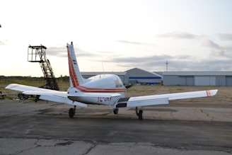 A small private airplane is parked on a tarmac near an aircraft hangar. The plane is white with red stripes and appears ready for takeoff or maintenance. The sky is overcast, and there's an aircraft ladder nearby.