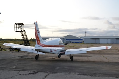 A small private airplane is parked on a tarmac near an aircraft hangar. The plane is white with red stripes and appears ready for takeoff or maintenance. The sky is overcast, and there's an aircraft ladder nearby.