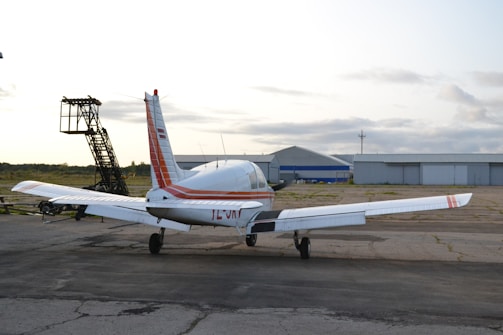 A small private airplane is parked on a tarmac near an aircraft hangar. The plane is white with red stripes and appears ready for takeoff or maintenance. The sky is overcast, and there's an aircraft ladder nearby.
