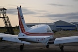 A small aircraft is parked on a tarmac near a hangar, with its tail and wings visible and marked with red and white stripes. The sky is overcast with clouds, and there's a structure with a staircase to the left.
