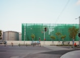 A construction site surrounded by a green netting with a large cylindrical tank on the left. Several small trees line the street in front, and there are traffic lights at the intersection. A person on a bicycle rides in the foreground near the crosswalk.