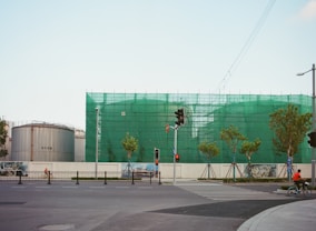A construction site surrounded by a green netting with a large cylindrical tank on the left. Several small trees line the street in front, and there are traffic lights at the intersection. A person on a bicycle rides in the foreground near the crosswalk.