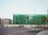 A construction site surrounded by a green netting with a large cylindrical tank on the left. Several small trees line the street in front, and there are traffic lights at the intersection. A person on a bicycle rides in the foreground near the crosswalk.