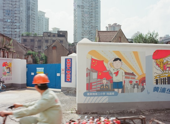 A street scene with construction barriers, one of which has colorful posters featuring cartoon-like illustrations, including a child with a red neckerchief saluting in front of Chinese flags and another poster on the side. There is a person in a construction helmet riding a bike in the foreground. The background consists of old buildings and tall modern apartment towers.