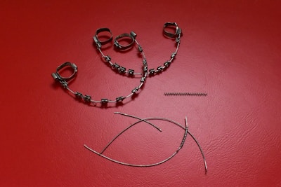 A collection of orthodontic appliance components laid out on a textured red surface. The items include metal braces with brackets and wire loops, a coil spring, and curved wires.