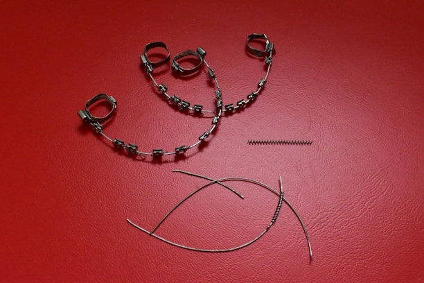 A collection of orthodontic appliance components laid out on a textured red surface. The items include metal braces with brackets and wire loops, a coil spring, and curved wires.