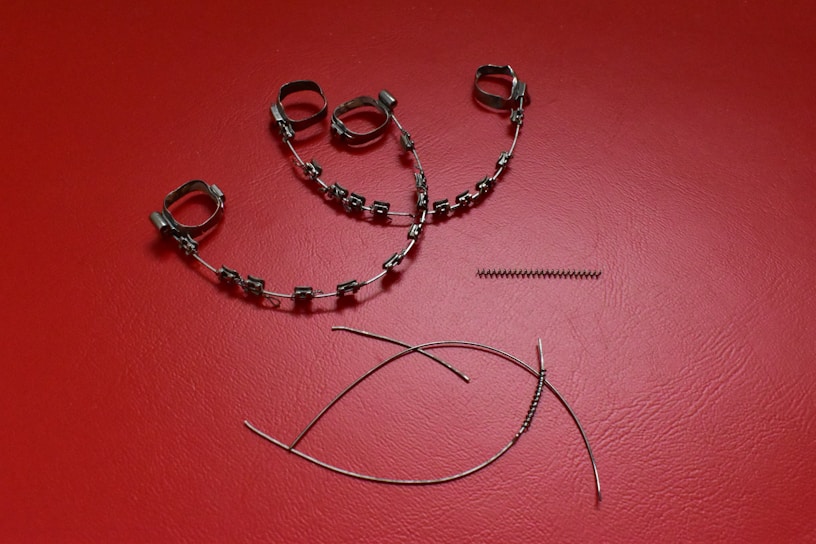 A collection of orthodontic appliance components laid out on a textured red surface. The items include metal braces with brackets and wire loops, a coil spring, and curved wires.