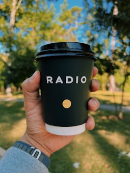 A hand is holding a black coffee cup with the word 'RADIO' printed on it in bold white letters and a small yellow dot underneath. The background features a sunny outdoor setting with trees and a clear blue sky.