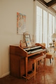 A wooden organ or piano is situated against a cream-colored wall, with a music sheet and microphone placed on top. Above it, there's a colorful artwork depicting a cartoon character in a top hat. To the right, a bright room is visible with a leather chair and a table with a lamp, receiving ample natural light through white window blinds.