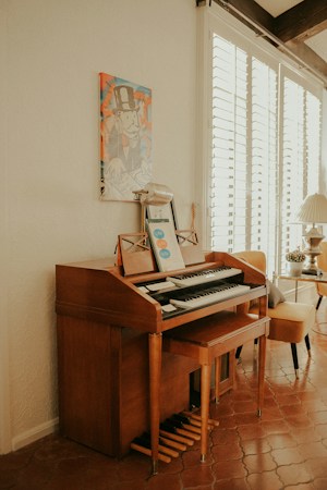 A wooden organ or piano is situated against a cream-colored wall, with a music sheet and microphone placed on top. Above it, there's a colorful artwork depicting a cartoon character in a top hat. To the right, a bright room is visible with a leather chair and a table with a lamp, receiving ample natural light through white window blinds.