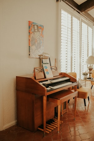 A wooden organ or piano is situated against a cream-colored wall, with a music sheet and microphone placed on top. Above it, there's a colorful artwork depicting a cartoon character in a top hat. To the right, a bright room is visible with a leather chair and a table with a lamp, receiving ample natural light through white window blinds.
