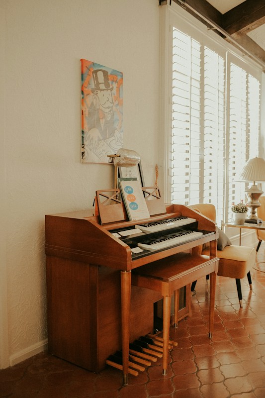 A wooden organ or piano is situated against a cream-colored wall, with a music sheet and microphone placed on top. Above it, there's a colorful artwork depicting a cartoon character in a top hat. To the right, a bright room is visible with a leather chair and a table with a lamp, receiving ample natural light through white window blinds.