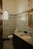 A bathroom with intricate, multicolored patterned tiles covering the walls and floor. The room features a sink with a wooden-framed mirror above it and a round window with a decorative pattern.
